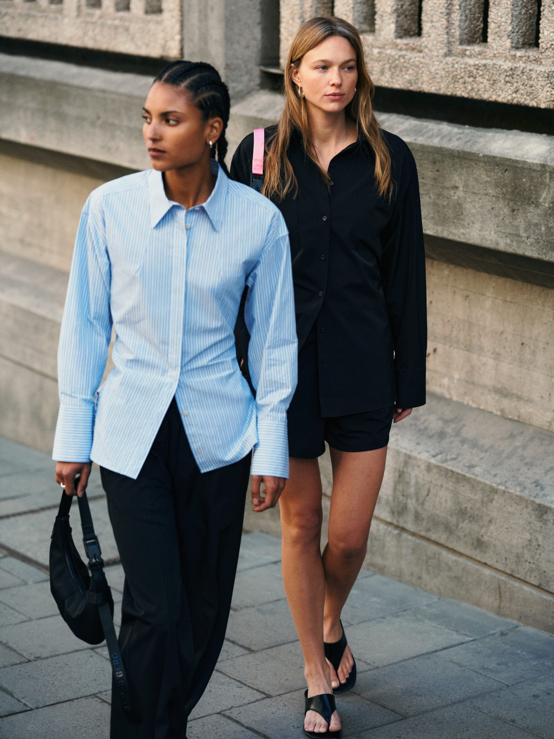 Two women walking outdoors on a city street, one in a light blue shirt and black pants, the other in a black shirt and shorts.