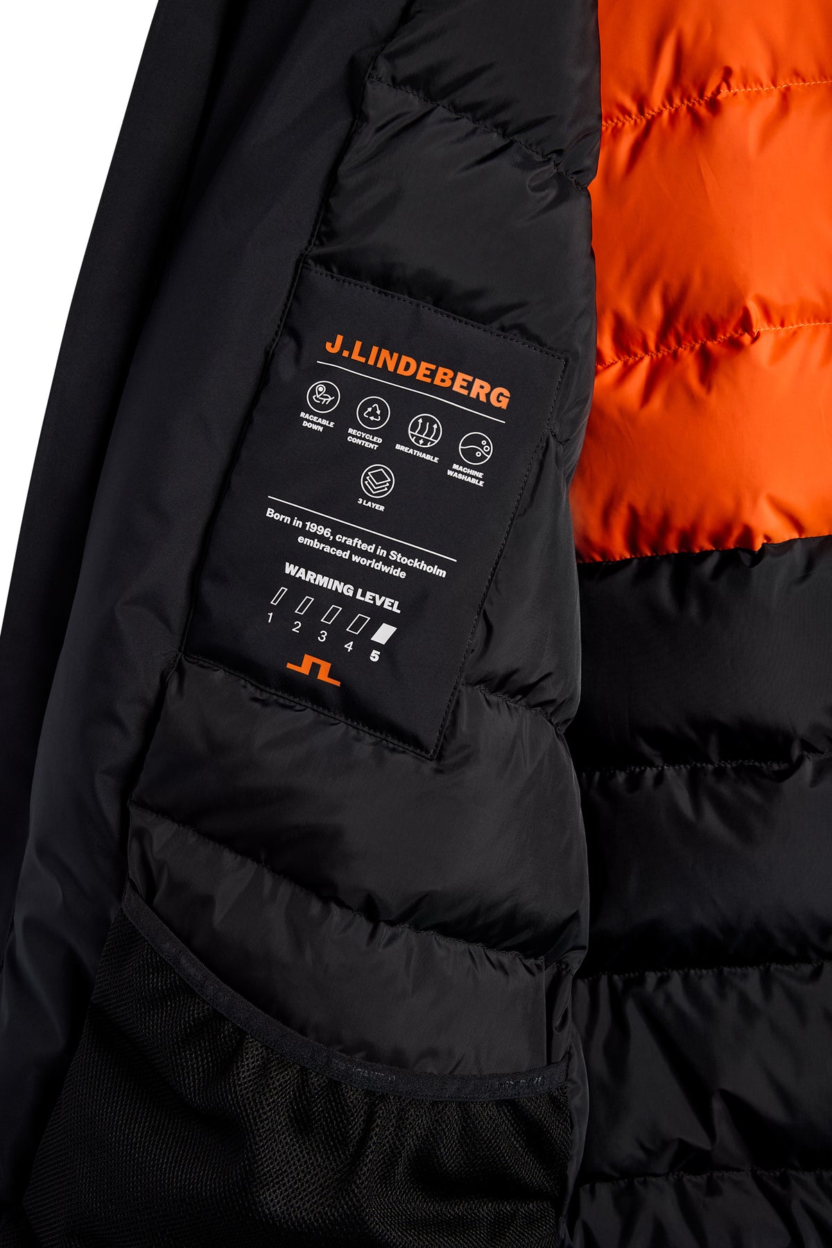 Close-up: Binnenzijde van de zwarte Alon Down Parka met gewatteerde zwarte en oranje voering en merklabel.