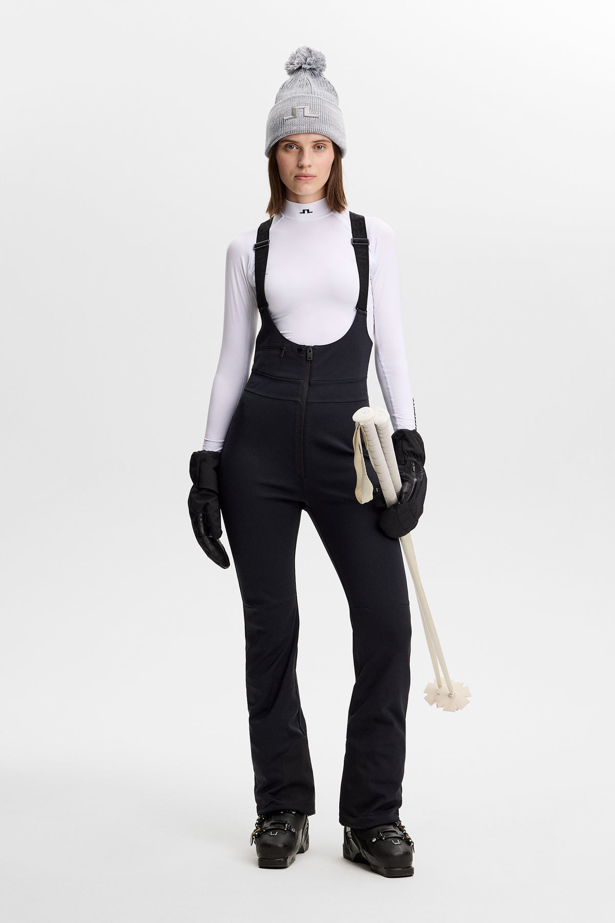 Vooraanzicht: Model draagt de zwarte Aviana Softshell Bib Pant met bretels, een witte top en skistokken.