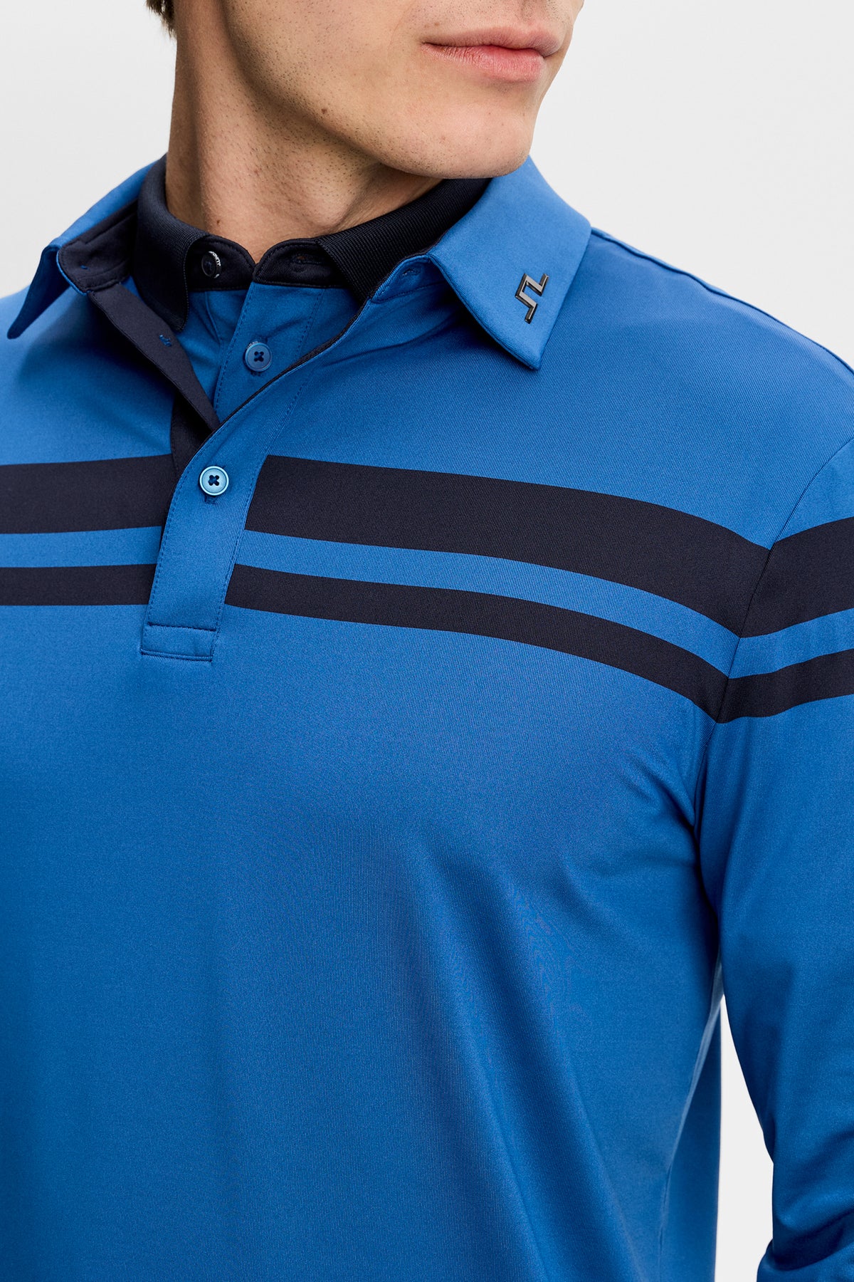 Nærbillede forfra: Blå Kenny LS stripe polo med mørke bryststriber og Bridge-logo på kraven.