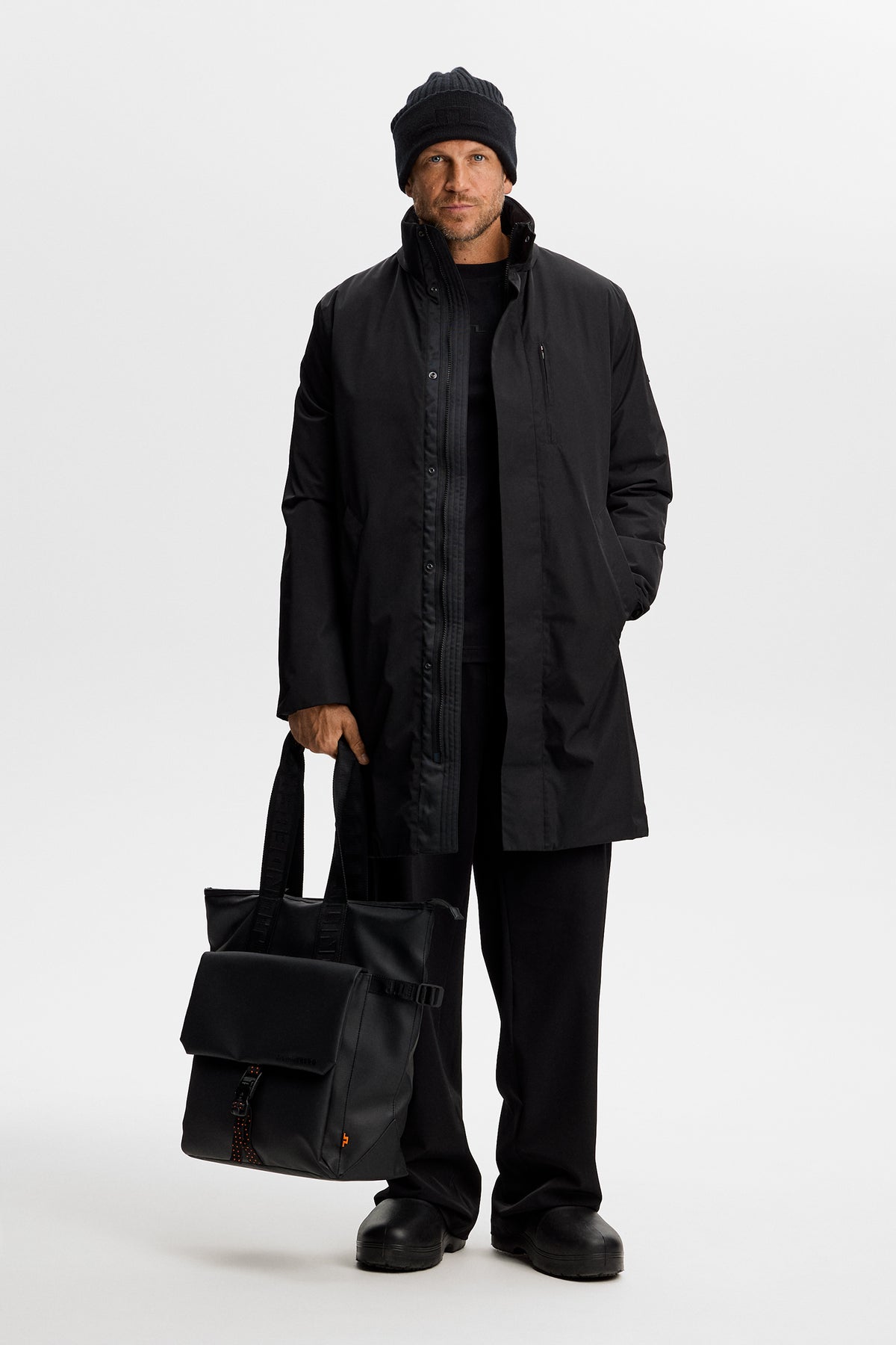 Model wearing the black Harold Padded Jacket unzipped over a dark outfit, holding a black bag in a studio setting.
