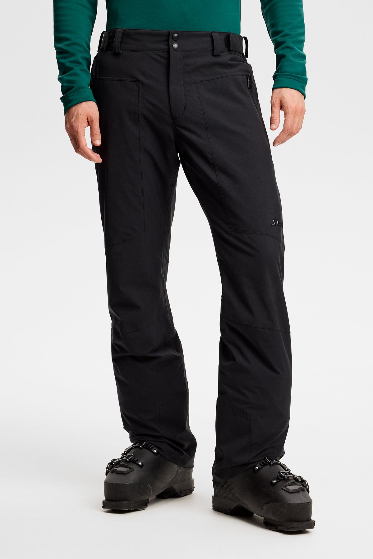 Vorderansicht: Schwarze Clarke Pant an einem Model, kombiniert mit schwarzen Skischuhen und Bridge-Logo am Bein.
