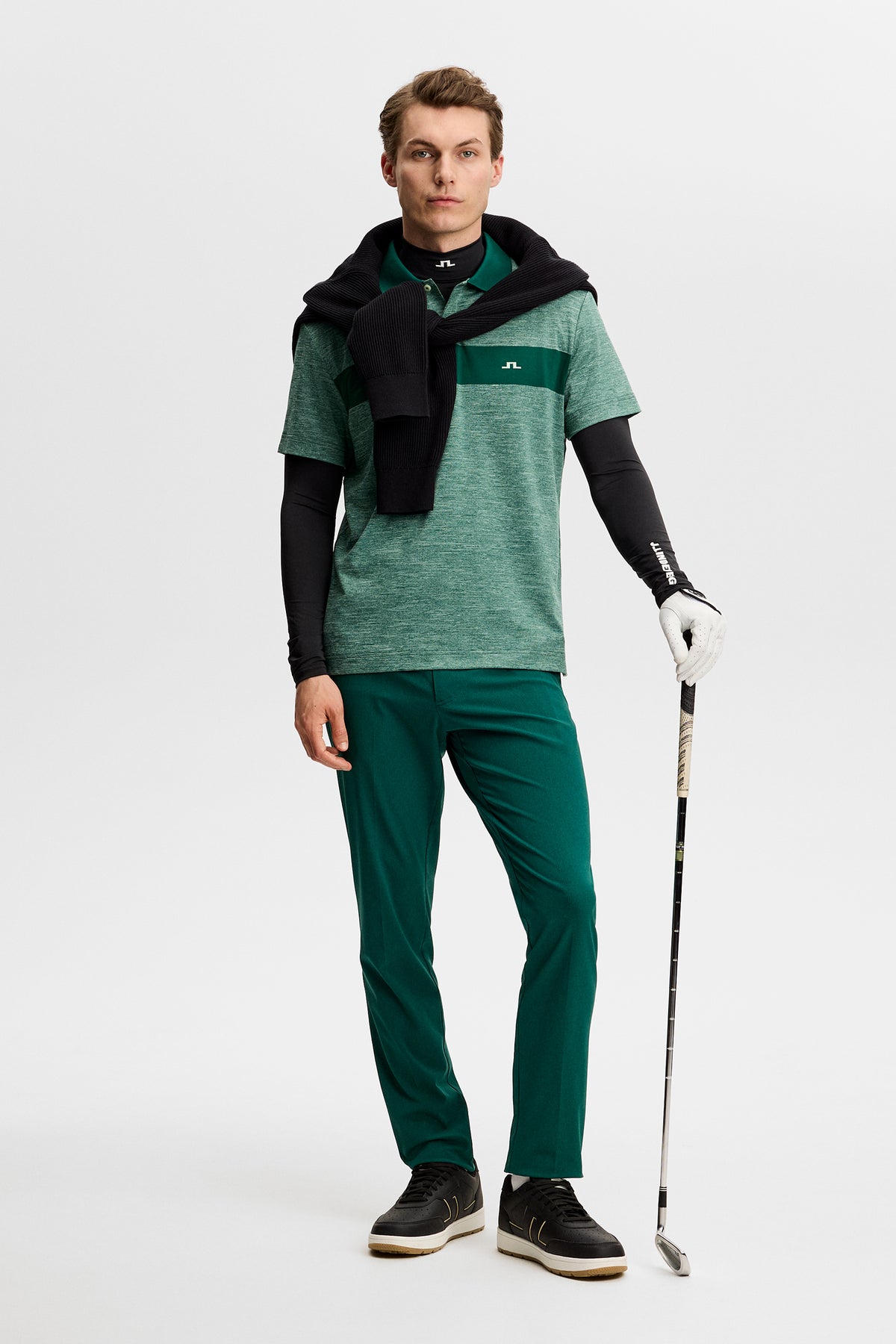 Model wearing the green melange Rocky Polo with a dark green chest stripe and Bridge logo, holding a golf club in a studio.