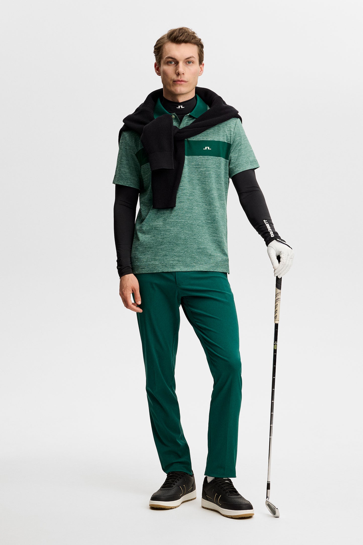 Model wearing the green melange Rocky Polo with a dark green chest stripe and Bridge logo, holding a golf club in a studio.