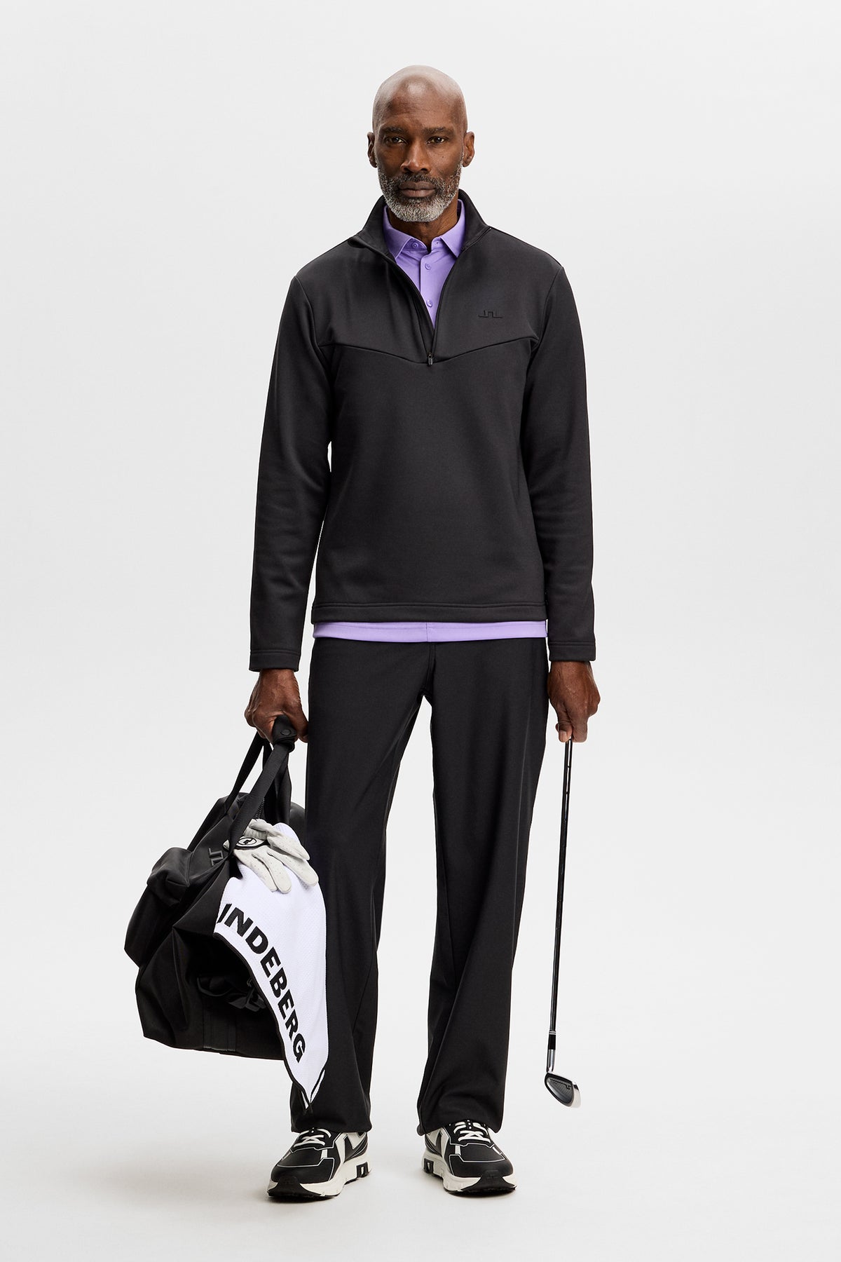 Helkropp framifrån: Modell bär Maclean Quarter Zip Mid Layer i Black med lila piké, svarta byxor och golfbag.