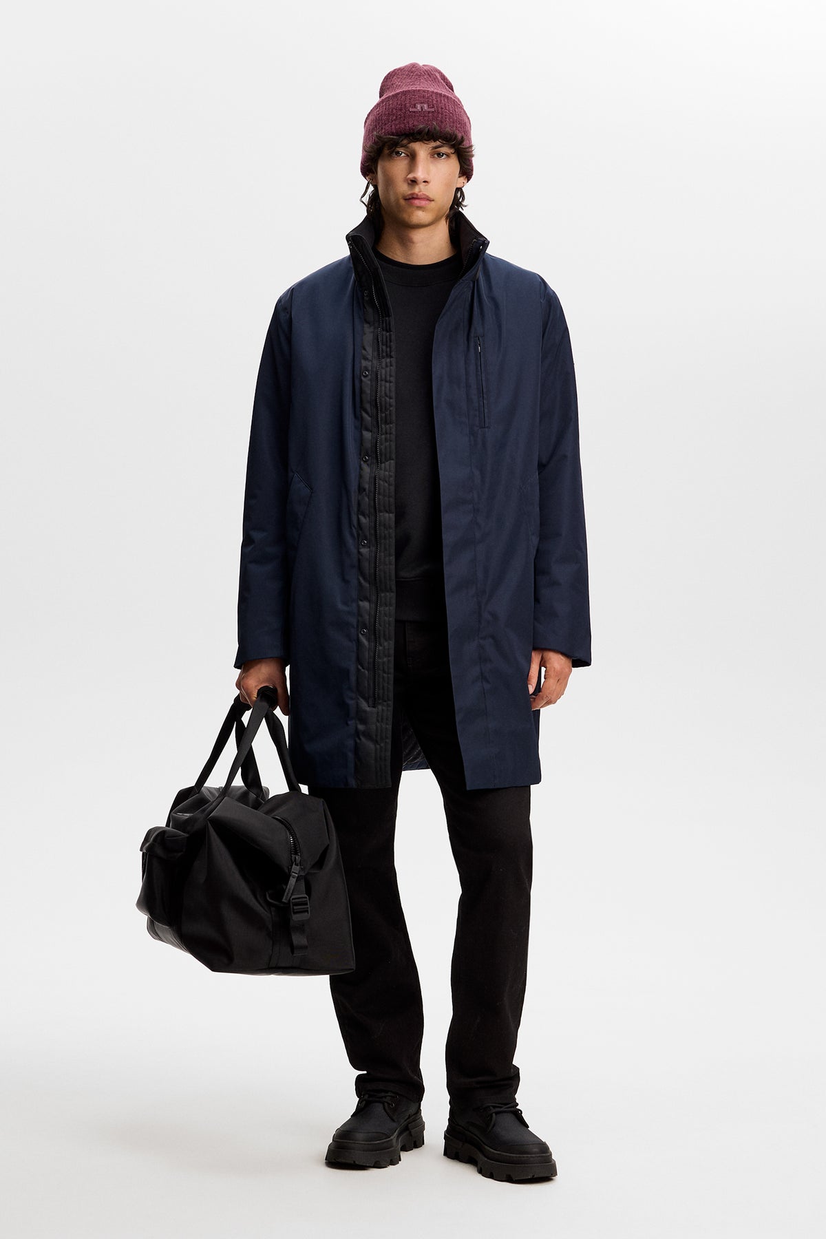 Model wearing the JL Navy Harold Padded Jacket open over a black outfit, holding a black bag in a studio setting.