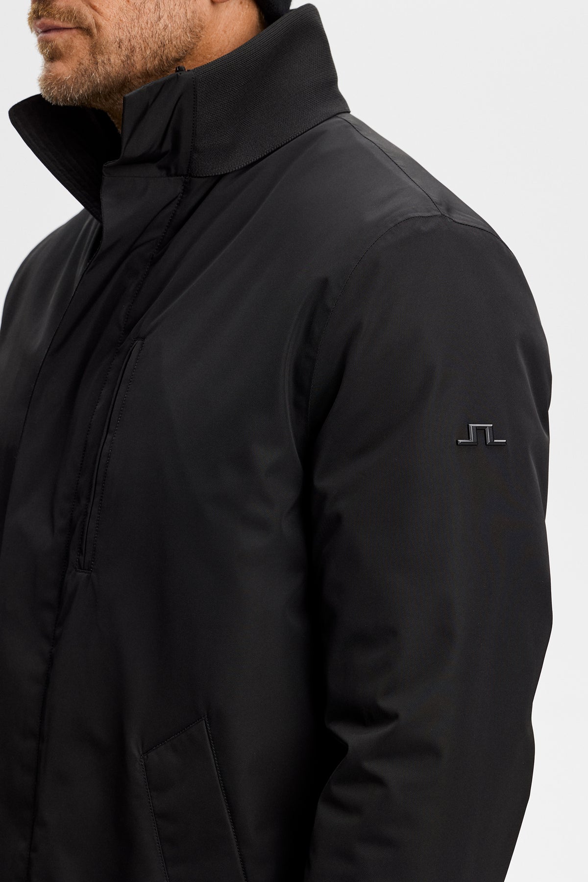 Close-up: Black Harold Padded Jacket showing the ribbed collar, chest pocket, and Bridge logo on the left sleeve.