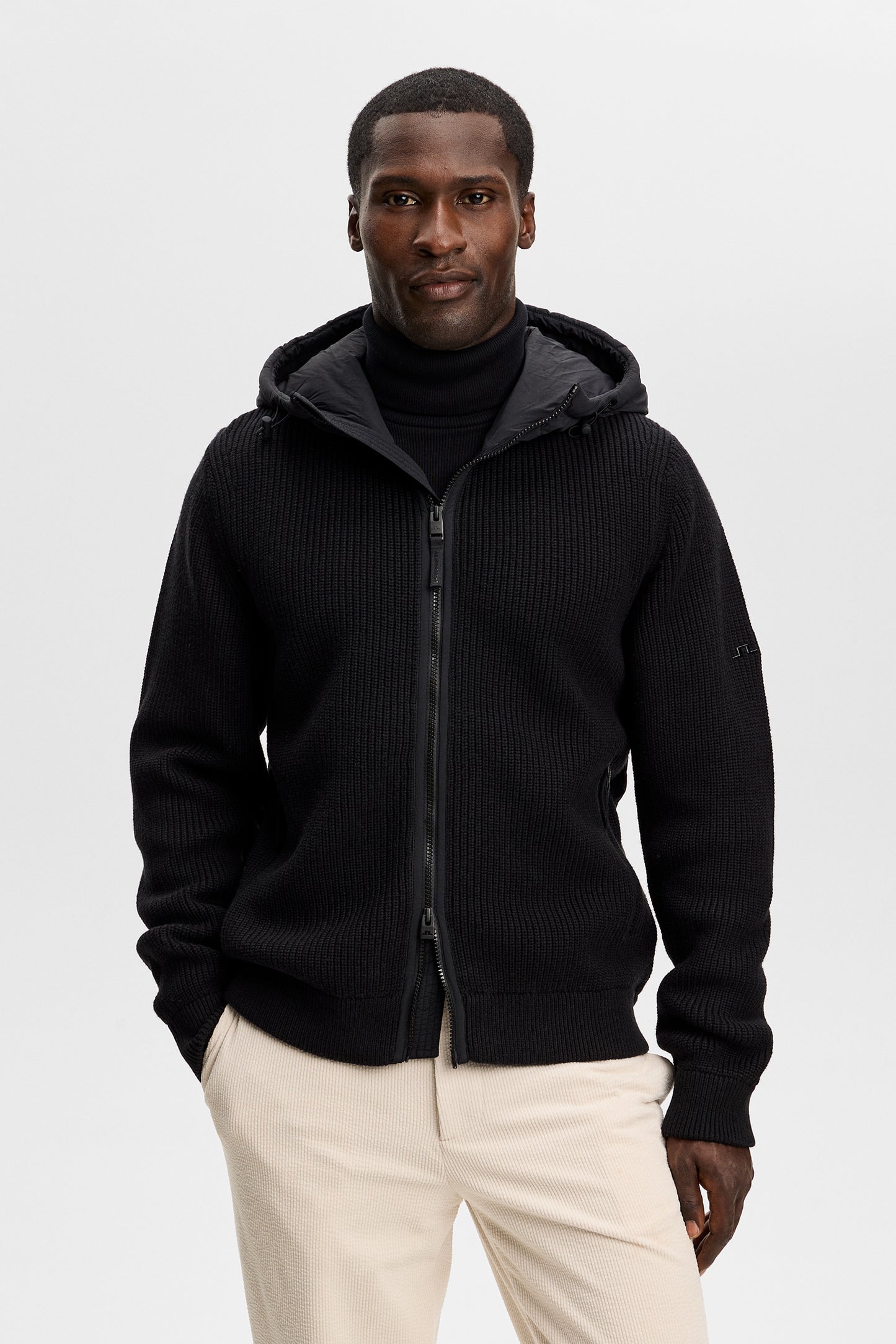 Framifrån: Modell bär Dorin Knit Hood Hybrid Jacket i svart med huva, tvåvägsdragkedja och Bridge-logotyp.