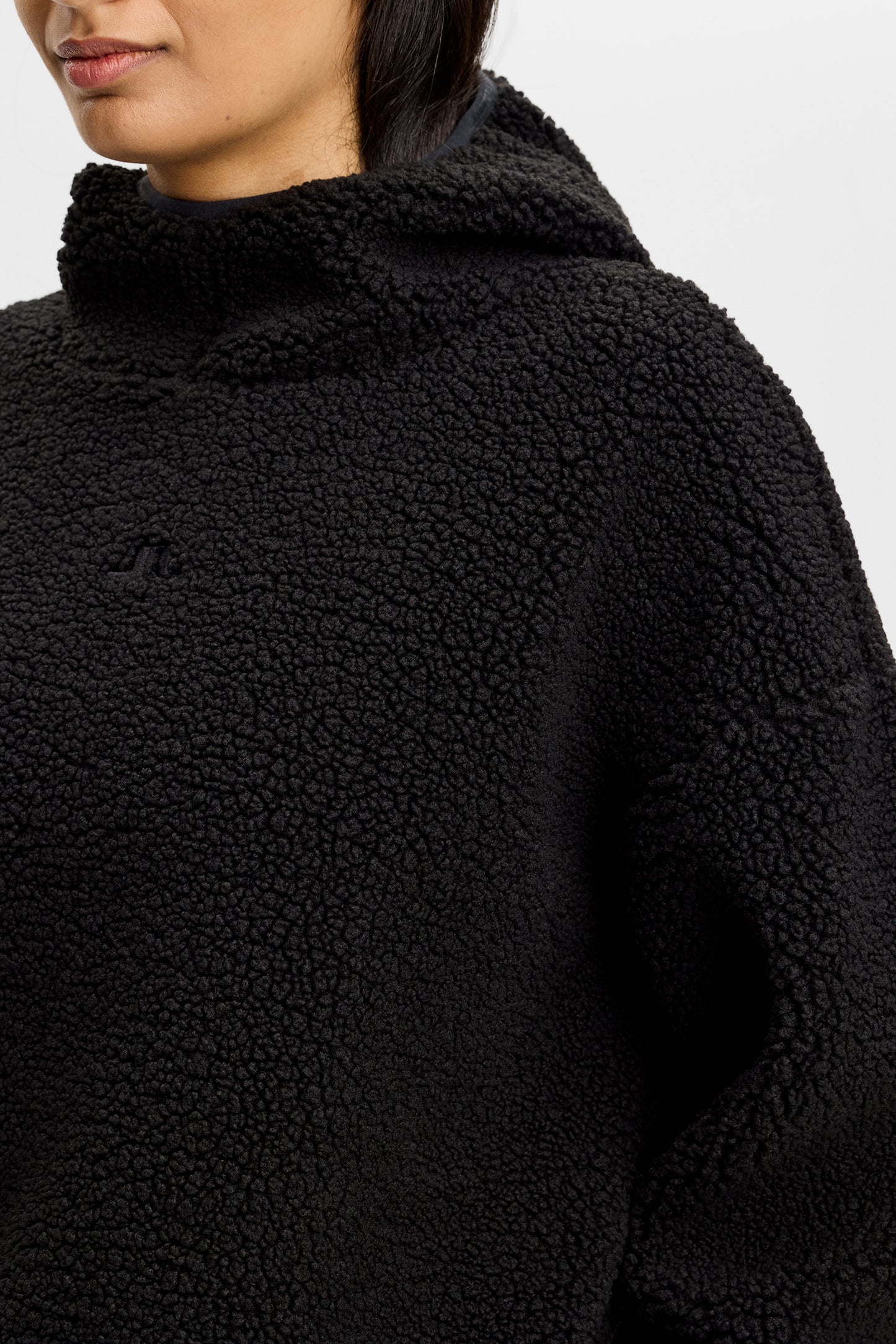 Detailansicht: Schwarzer Comona Pile Hood aus Teddy-Fleece mit fester Kapuze und tonalem Bridge-Logo auf der Brust.