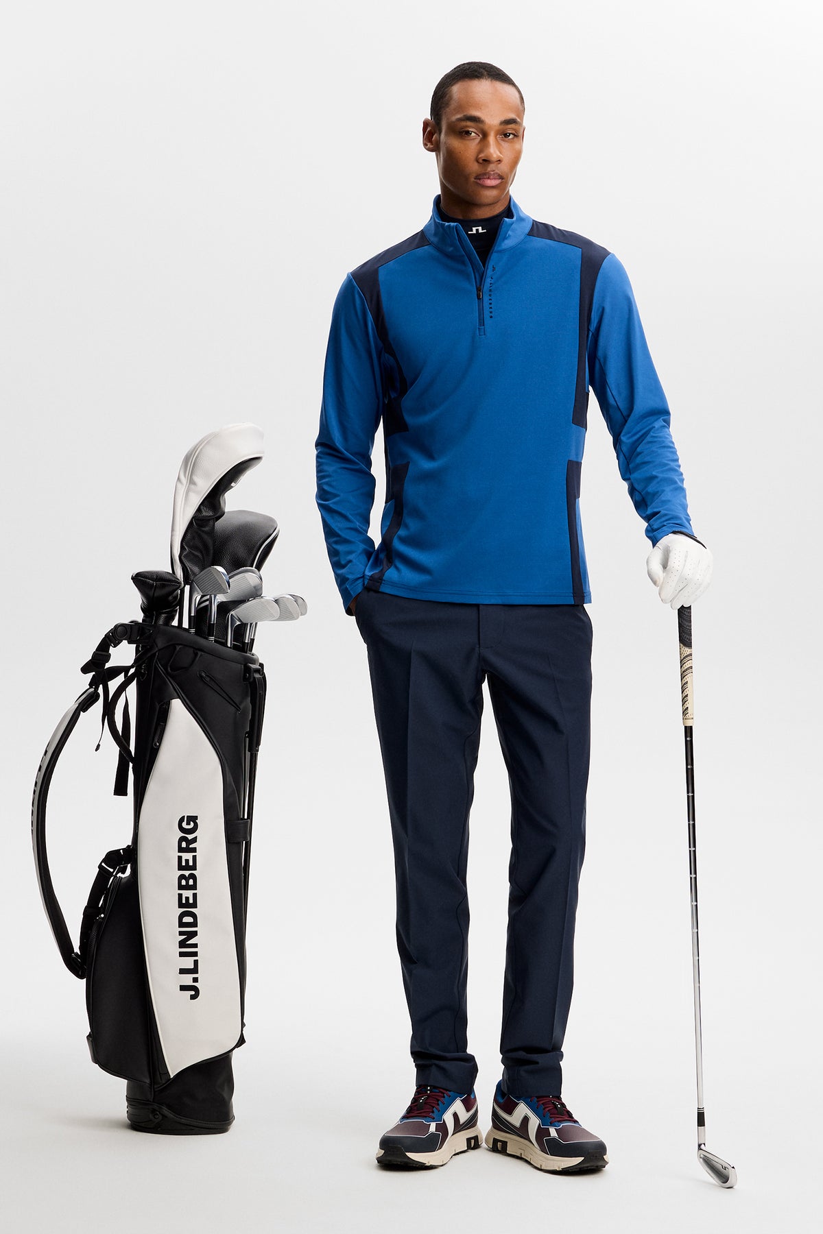 Vooraanzicht: Model draagt de blauwe Finn Quarter Zip Mid Layer met color-block panelen, donkere broek en golftas.