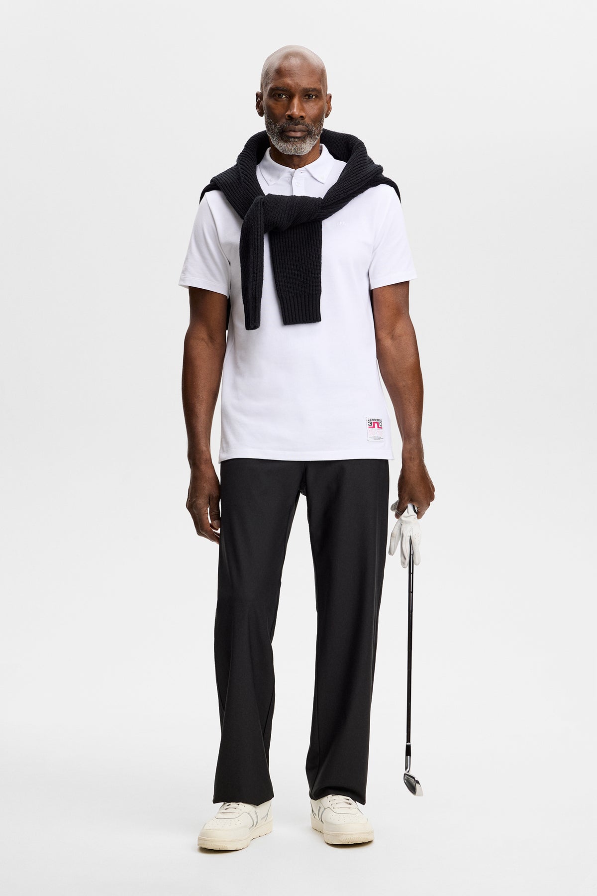 Vooraanzicht: Model draagt de Verse Button Down Polo in White met zwarte trui om de schouders, zwarte broek en golfclub.