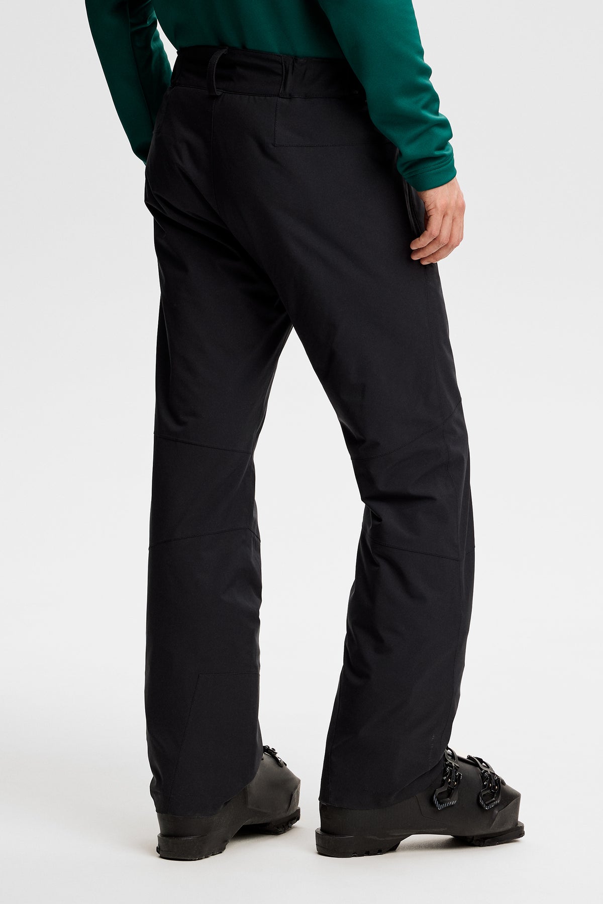 Rückansicht: Clarke Pant in Schwarz an einem Model, kombiniert mit schwarzen Skischuhen.