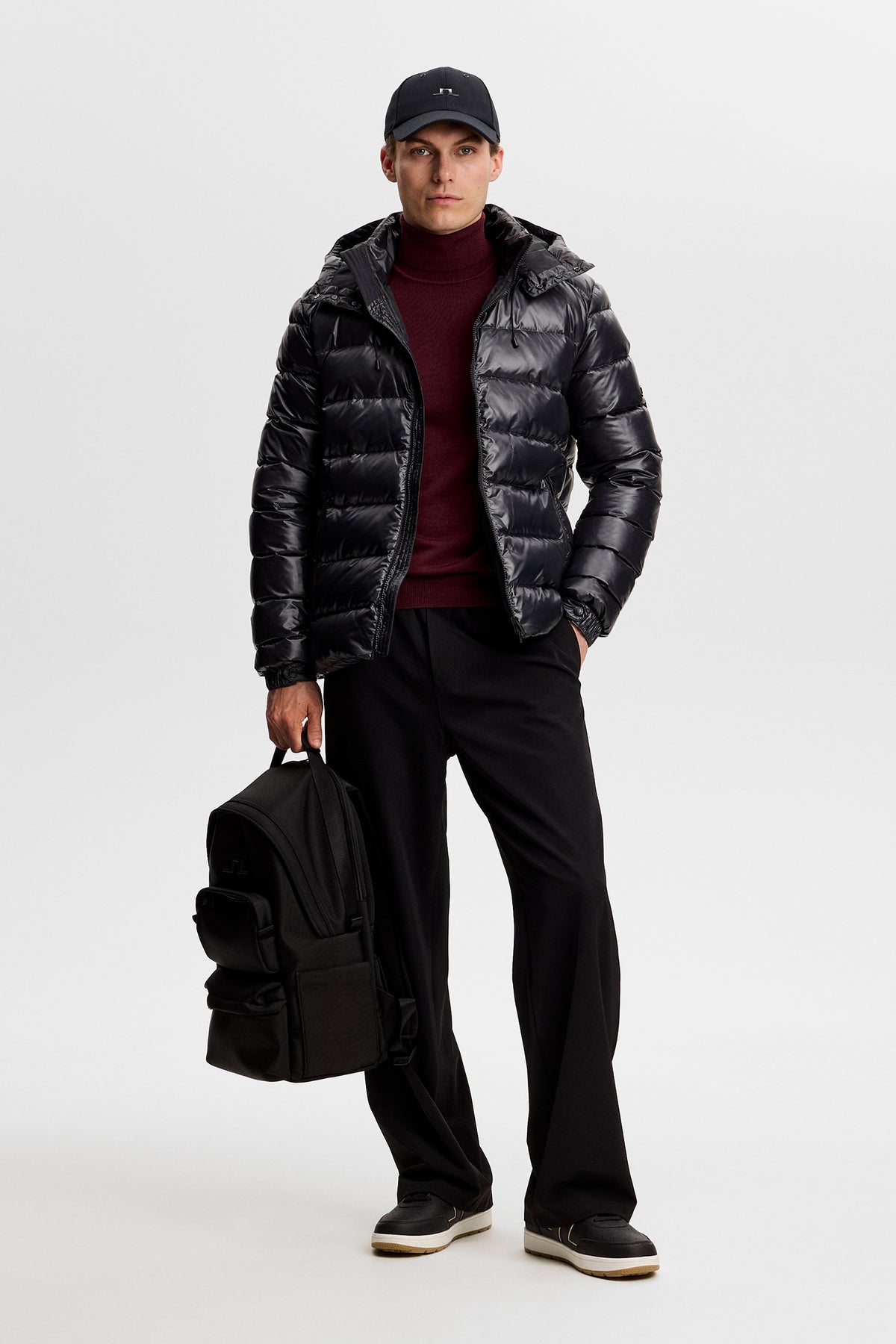 Vorderansicht: Schwarze Creek Light Down Jacket an einem Model mit dunkelrotem Rollkragenpullover und schwarzem Rucksack.