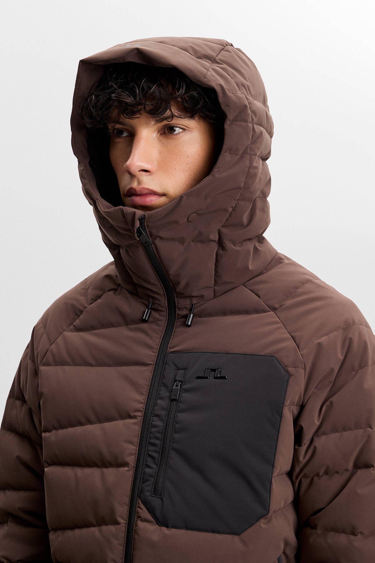 Thornton Down Jacket in Seal Brown, close-up with raised hood and black zippered chest pocket with Bridge logo.