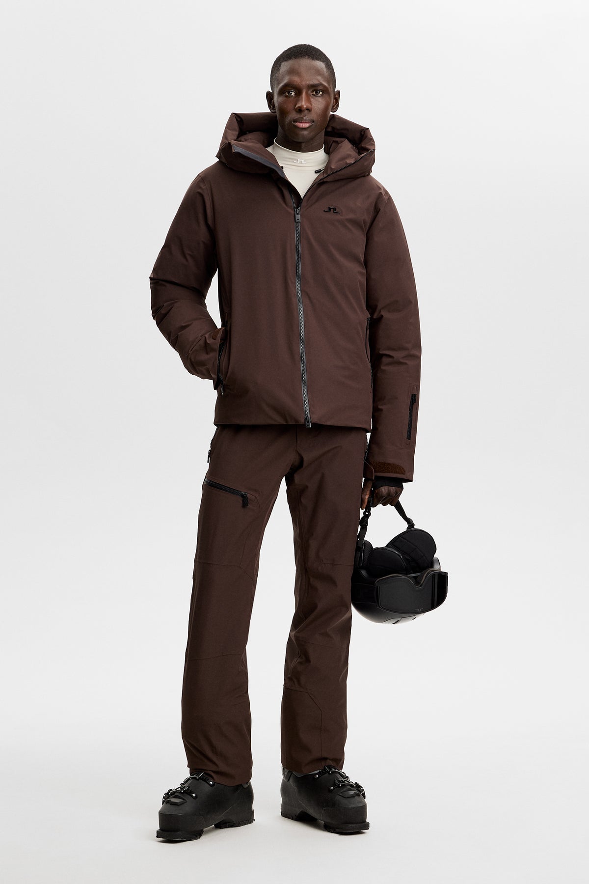 Forfra: Crue Suspender Pant i Seal Brown, båret med matchende skijakke, skihjelm og skistøvler.