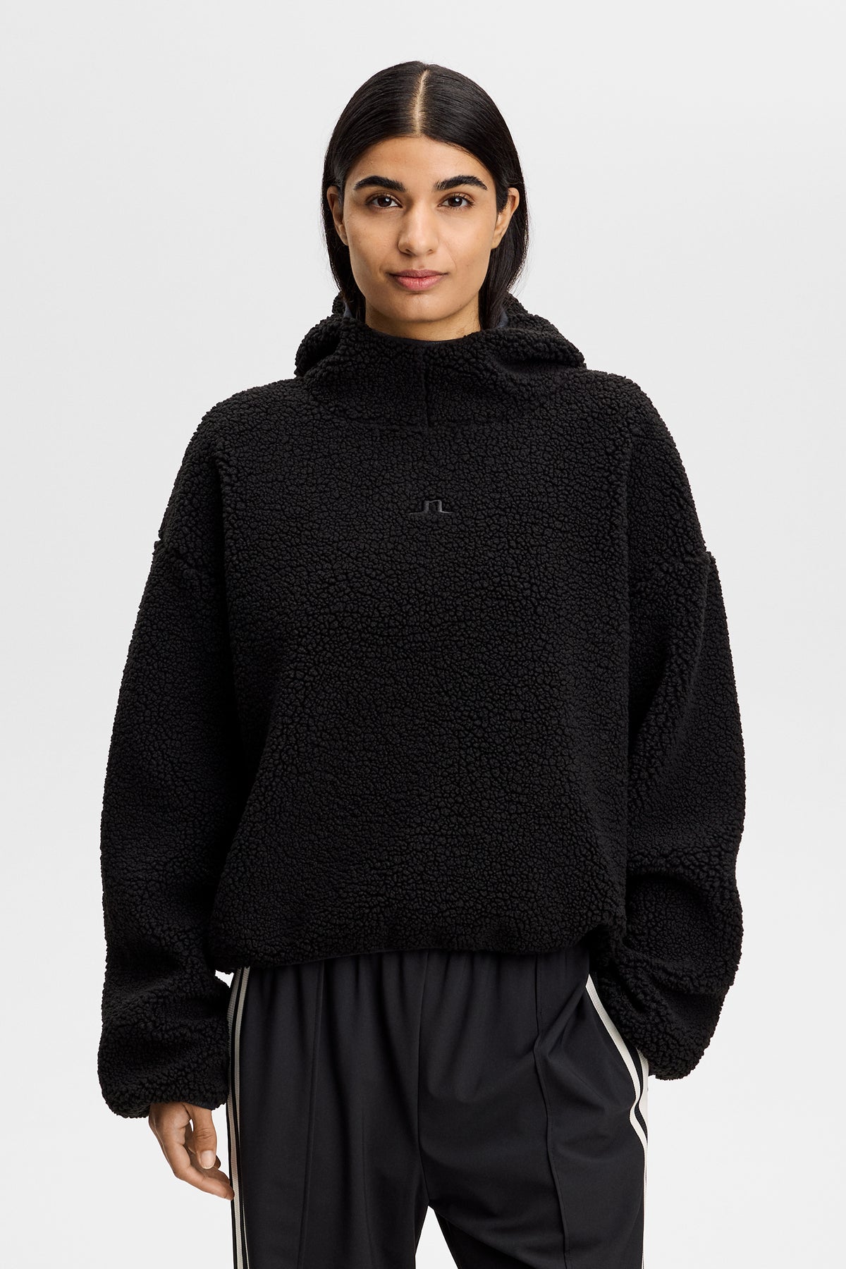 Vorderansicht: Schwarzer Comona Pile Hood aus Teddy-Fleece mit Kapuze und Bridge-Logo auf der Brust.
