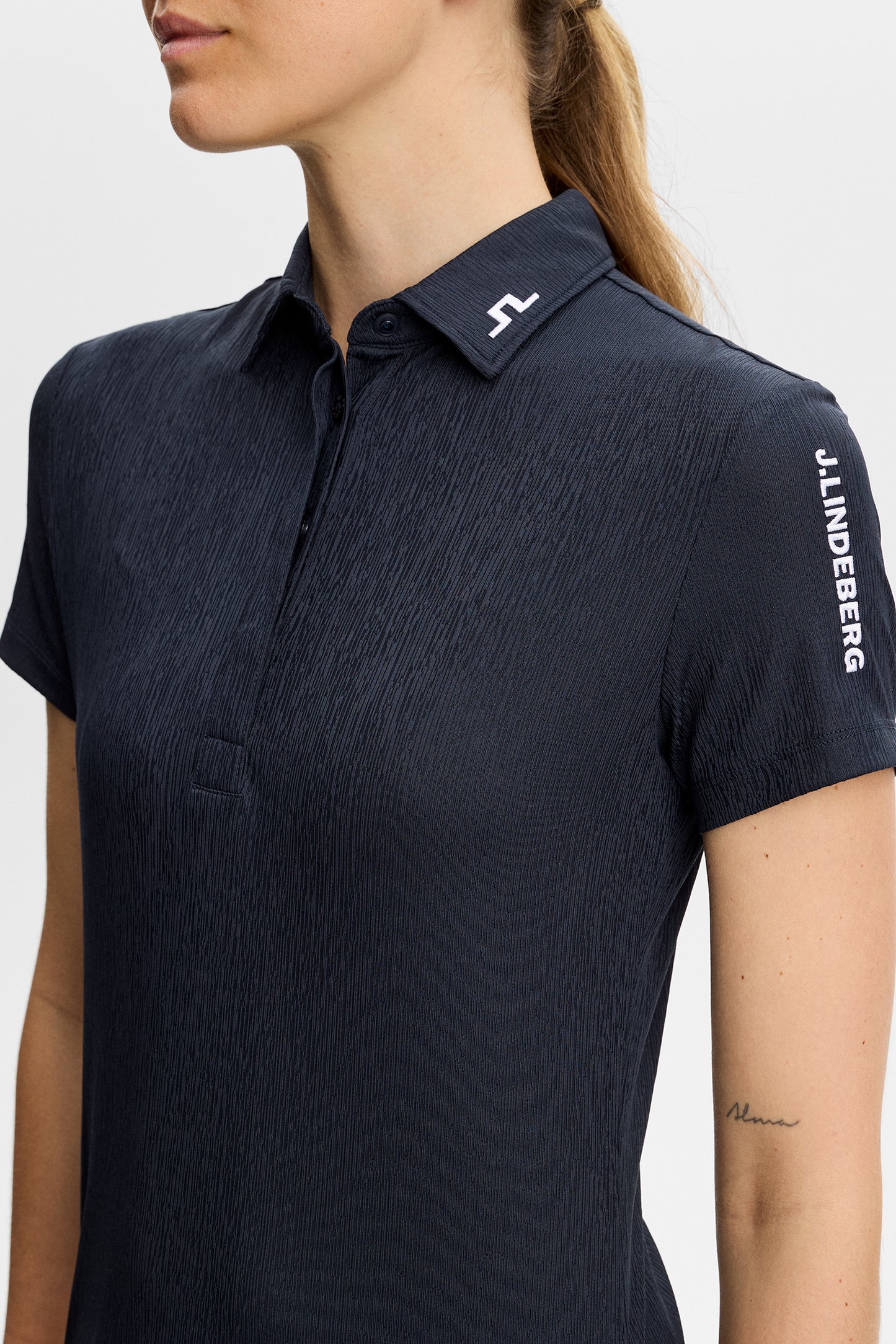 Detailansicht: Tour Tech Textured Polo in JL Navy mit Bridge-Logo am Kragen und J.Lindeberg-Schriftzug am Ärmel.
