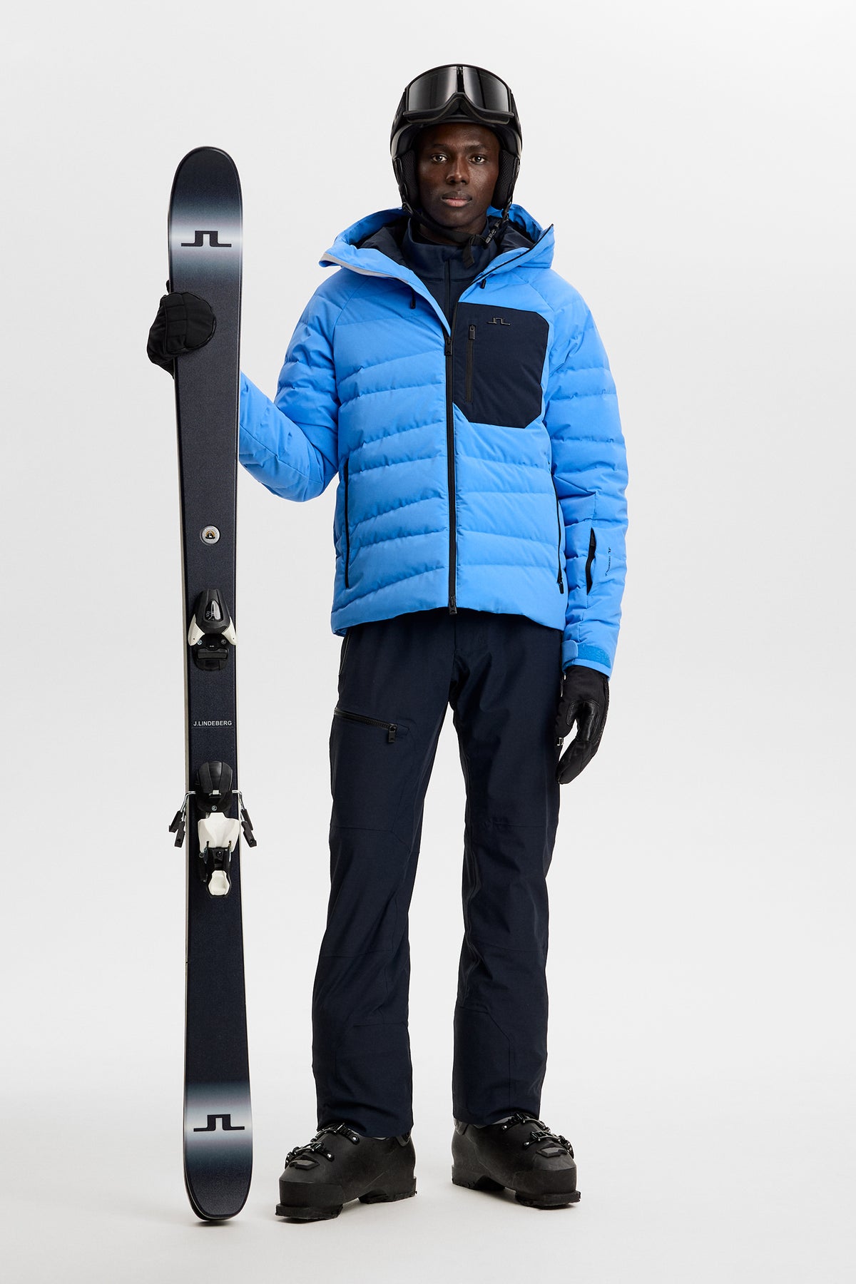 Full-body view: Model wearing the Thornton Down Jacket in Sonic Blue styled with ski pants and holding skis.
