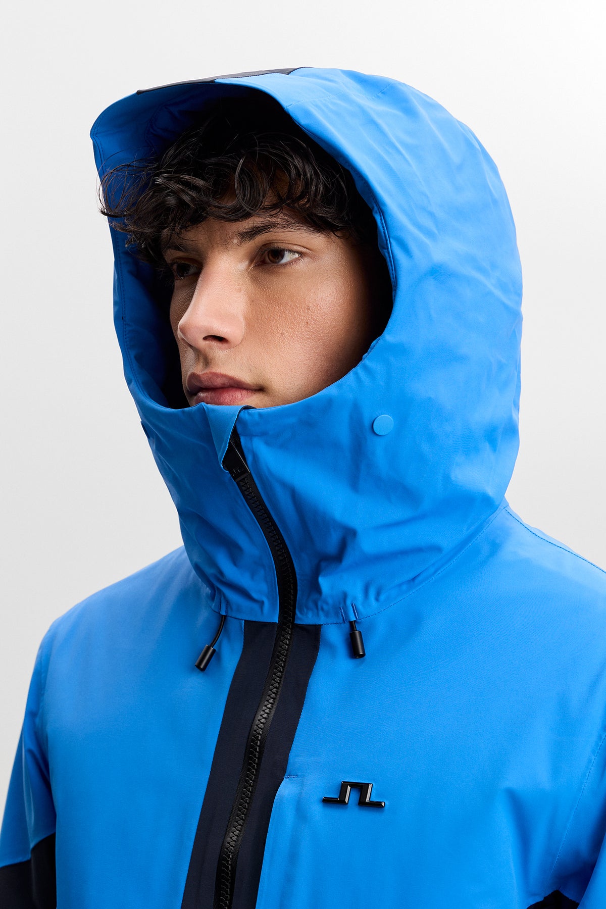 Detailansicht: Caldwell Jacket in Sonic Blue mit aufgesetzter Kapuze, hohem Kragen und Bridge-Logo auf der Brust.