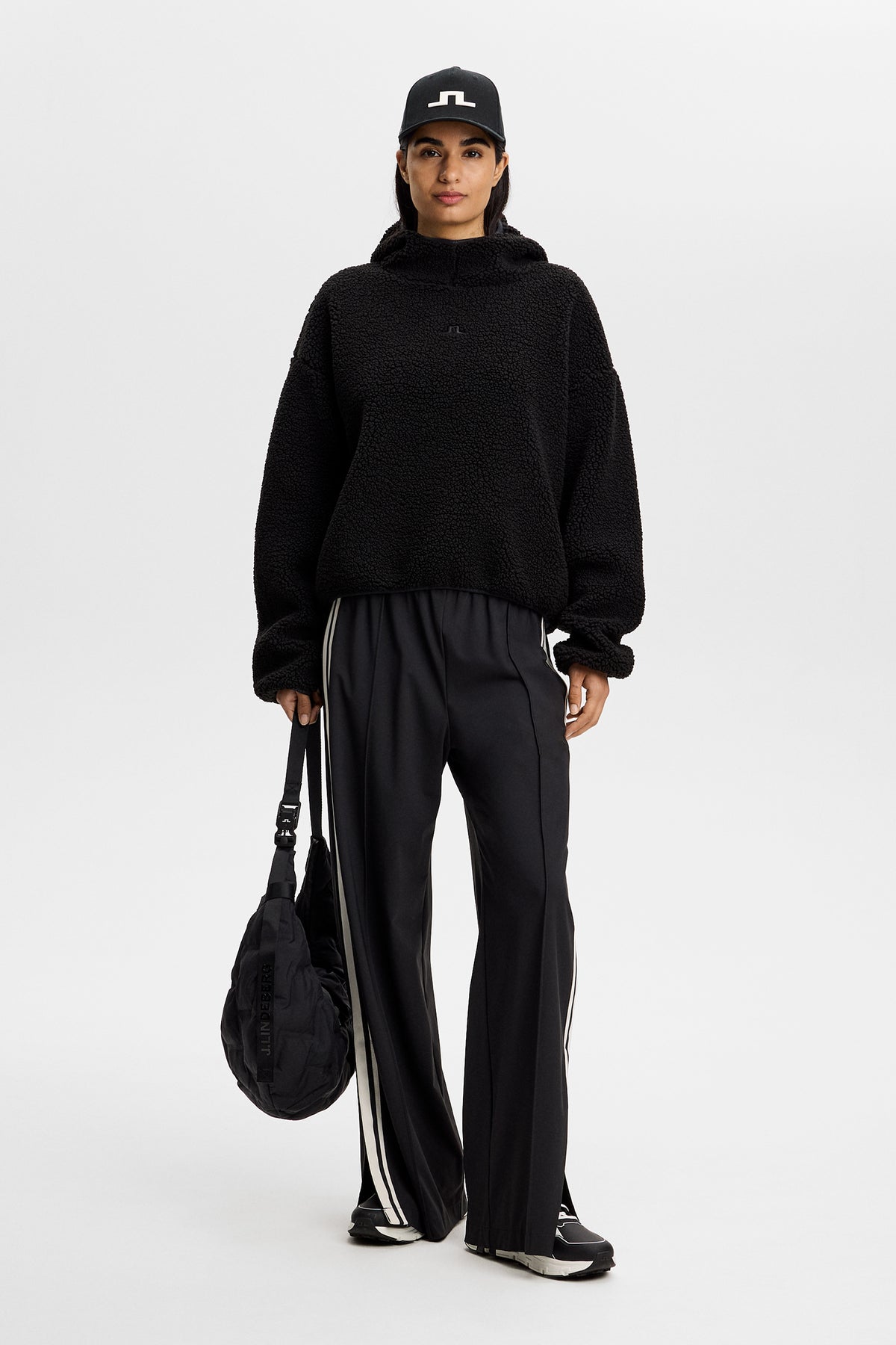 Vorderansicht: Schwarzer Comona Pile Hood aus Teddy-Fleece an einem Model mit schwarzer Hose, Cap und Tasche.