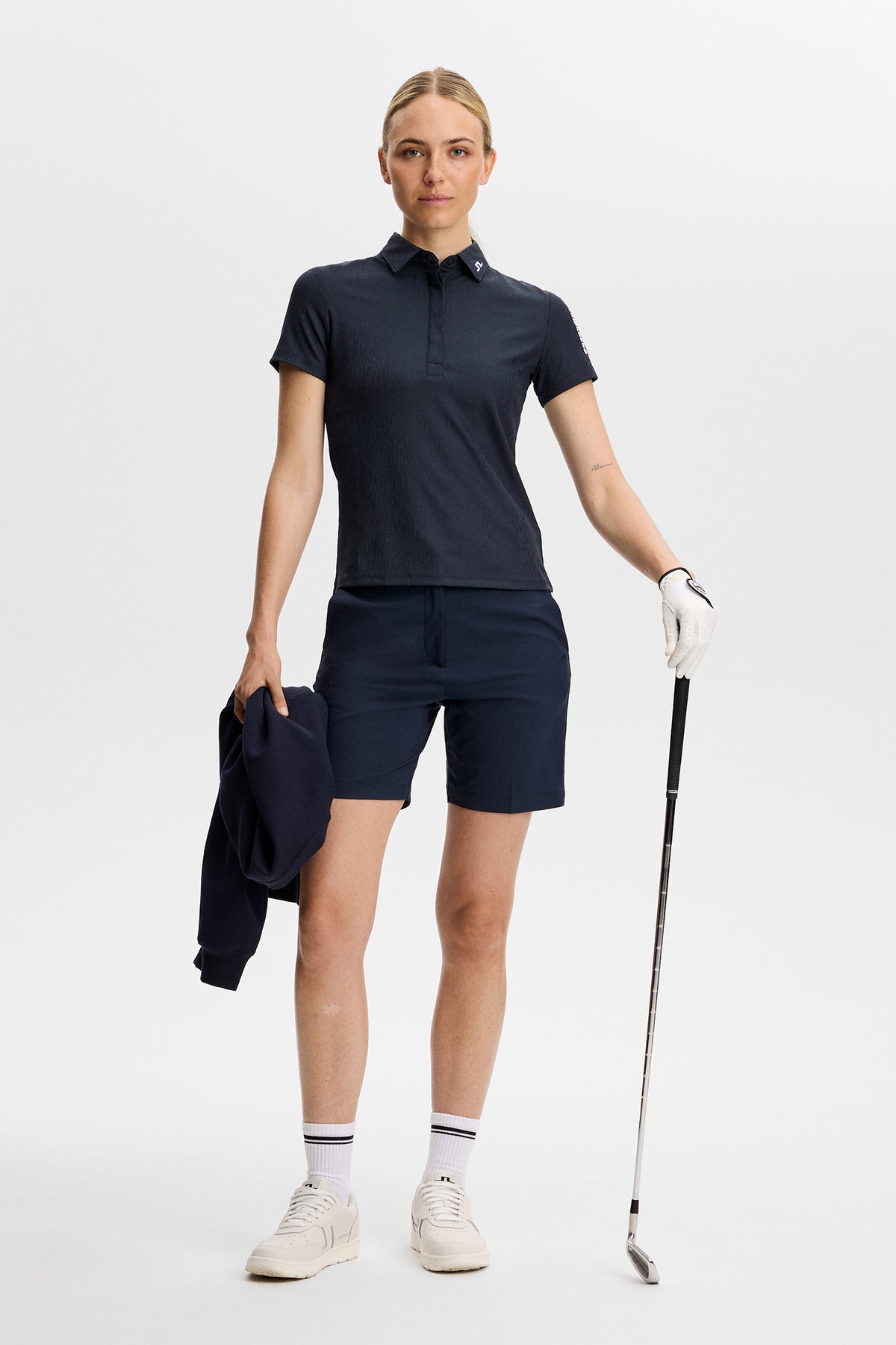 Vorderansicht: Tour Tech Textured Polo in JL Navy an einem Model, kombiniert mit passenden Shorts und Golfschläger.