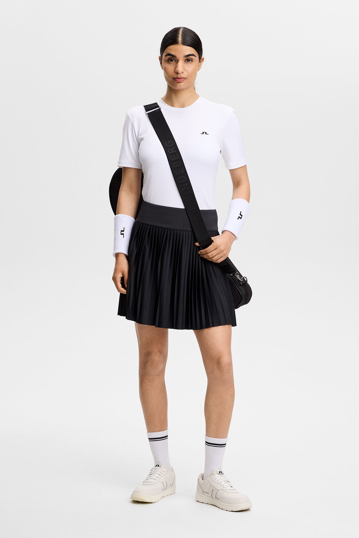 Vooraanzicht: Model draagt de zwarte geplooide Alexandra Skirt met een brede tailleband en wit T-shirt.