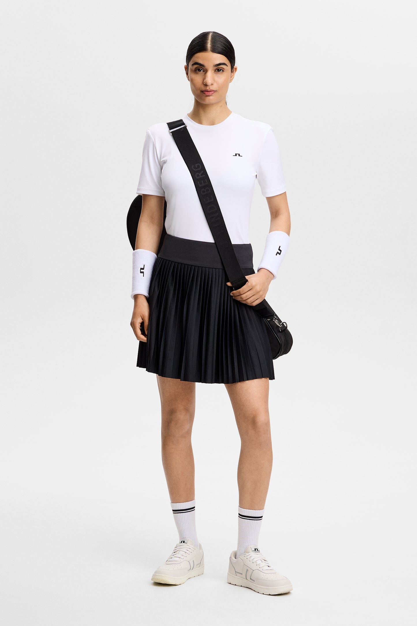 Vooraanzicht: Model draagt de zwarte geplooide Alexandra Skirt met een brede tailleband en wit T-shirt.