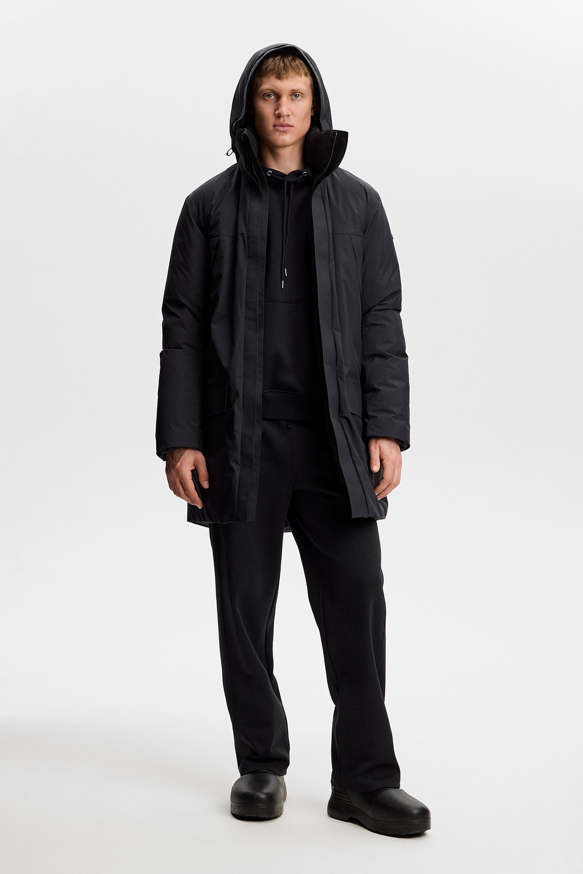 Vooraanzicht: Model draagt de zwarte Alon Down Parka met capuchon open over een zwarte hoodie en broek.